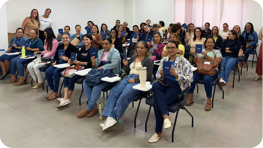 Turma de professoras em uma sala de formação, sorrindo e segurando cadernos azuis